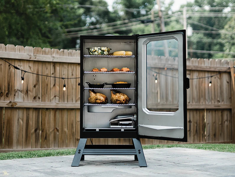 An open Digital Electric Smoker with a window in the door and legs to raise the smoker off the ground. Inside are a variety of vegetables, sides, planked salmon filets, and whole chickens. The water tray and side loading wood chip holder are in the bottom of the smoker.