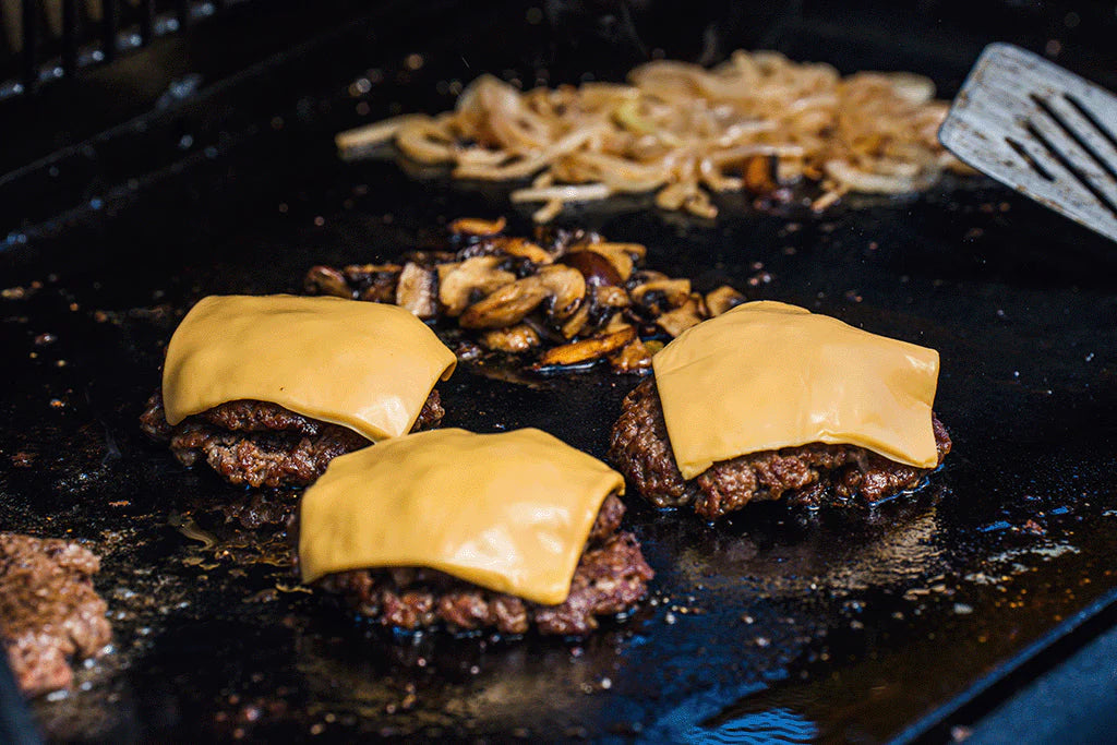 Three burgers with cheese cooking on a griddle while onions and mushrooms sauté in the background.
