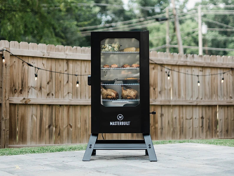 A Masterbuilt Digital Electric Smoker with legs and a window in the door sits on an outdoor patio. There is a wooden privacy fence strung with Edison bulks in the background. 4 racks of food including whole chickens, vegetables, and cast iron pans are visible through a window in the smoker door.