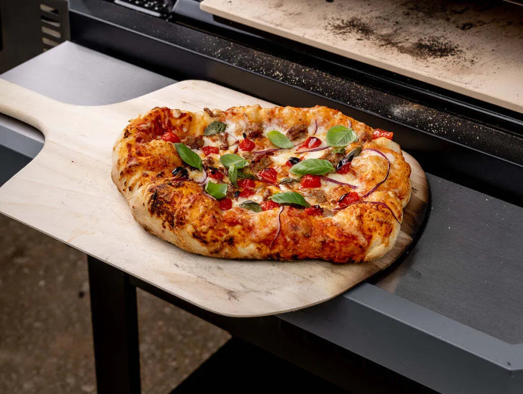 A backed Sausage Tomato Basil pizza rests on a pizza peel placed on the front shelf of a Gravity Series grill. The front of the pizza oven is behind the pizza.