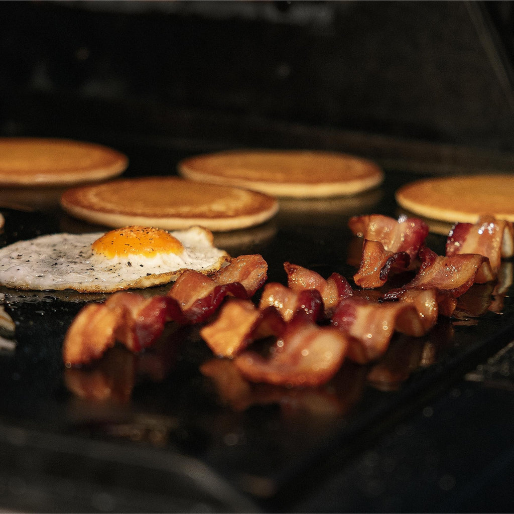 A seasoned egg, strips of bacon, and pancakes all cook on a griddle