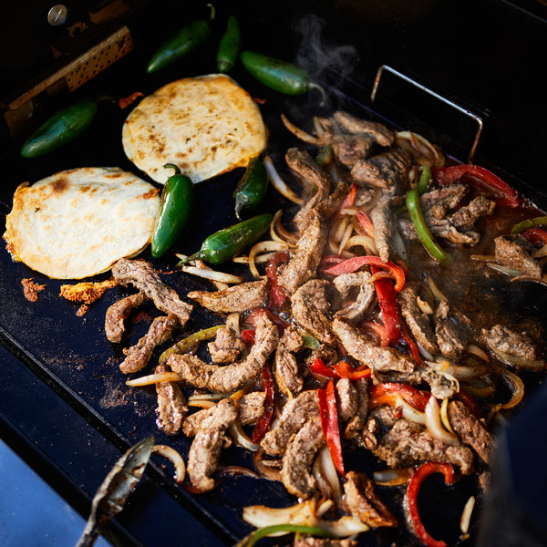Fajita meat with sliced bell peppers and onions finish cooking on a griddle. Whole jalapenos and tortillas grill to the side of the meat.